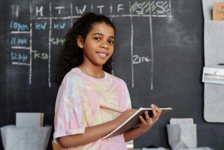 female student writing in a notebook in front of a classroom chalkboard